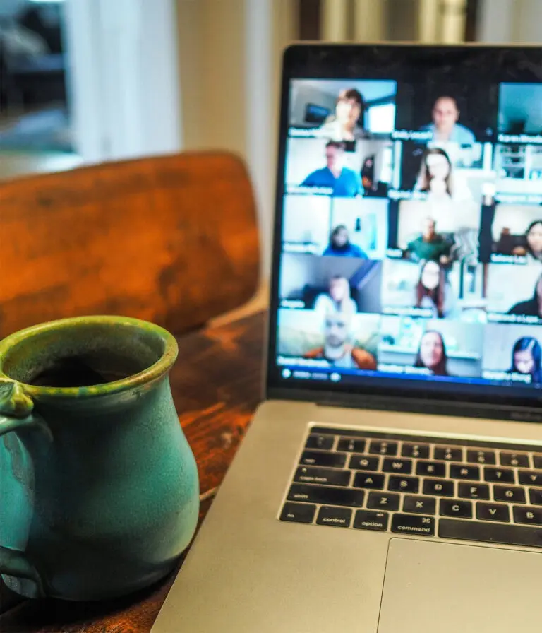 A laptop screen shows lots of small screens of people on a zoom call. A cup sits beside the laptop on the table.