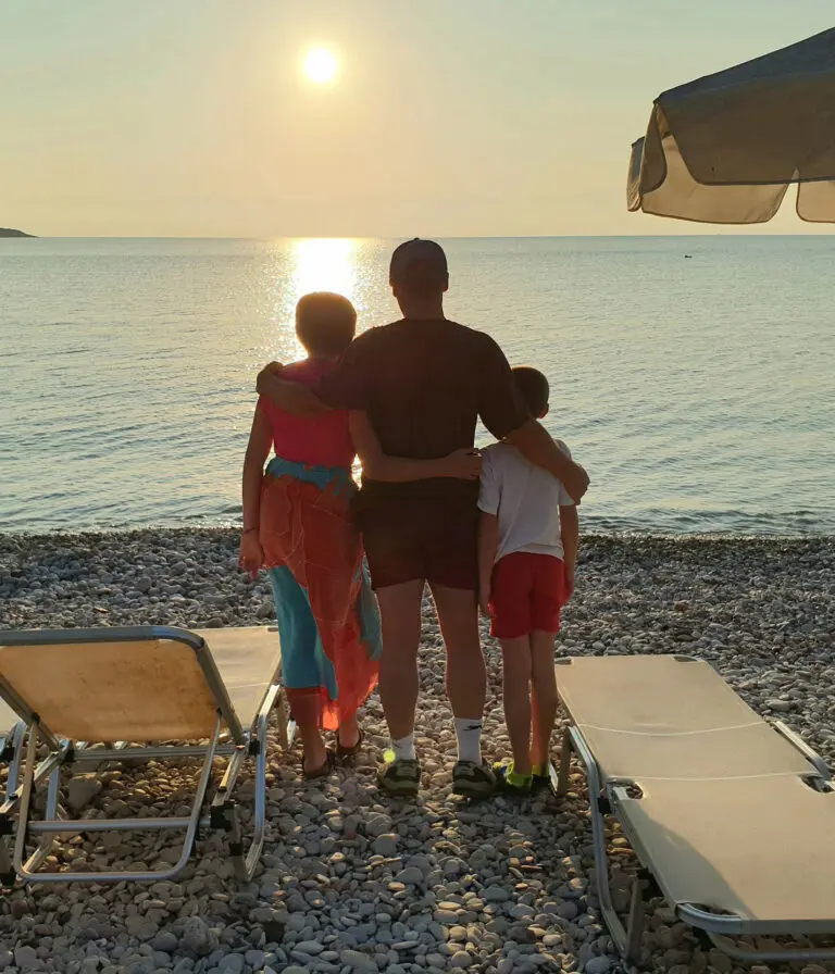 A father stands with his two children on a stoney beach looking out over the water at sunset with beach chairs beside them.
