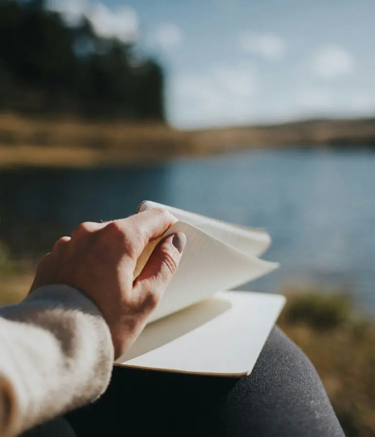 A close up of a woman flicking through a lined note pad whilst sitting at a lake that is blurred in the distance.