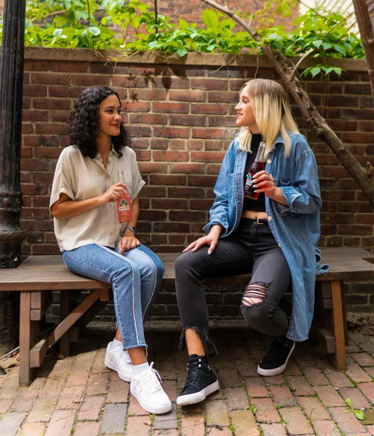 Two women chat, sitting on a wooden bench with a brick wall behind them.
