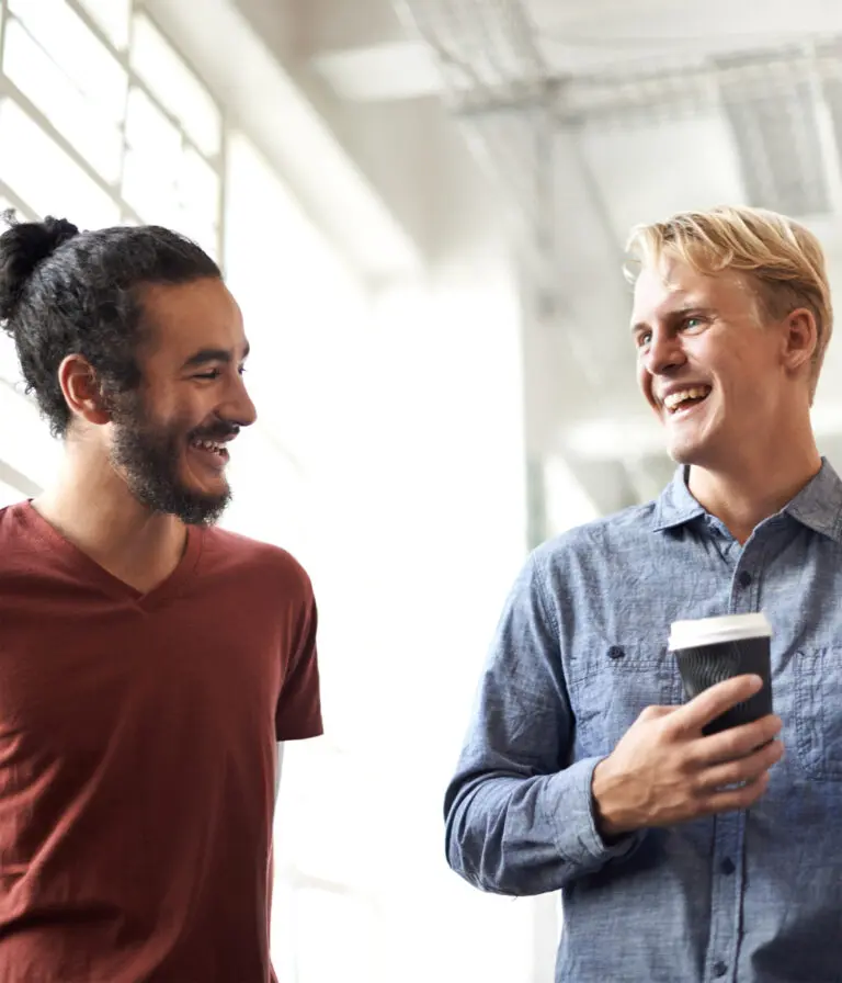 Two men chat and smile as one of them drinks from a take away coffee cup