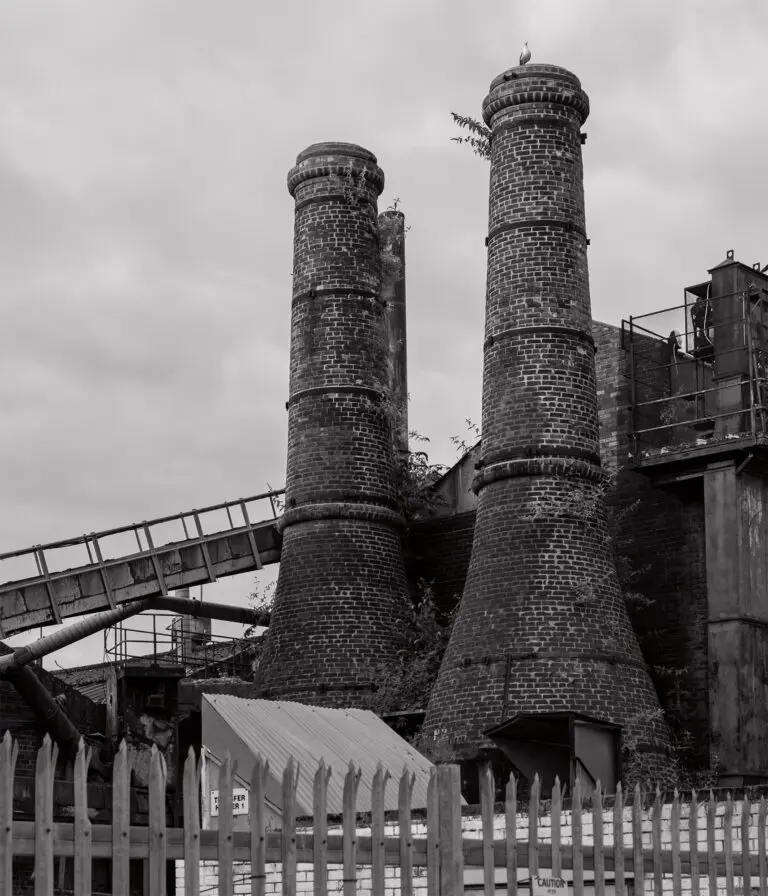 A low angle of a disused facility with stone chimneys covered in weeds.