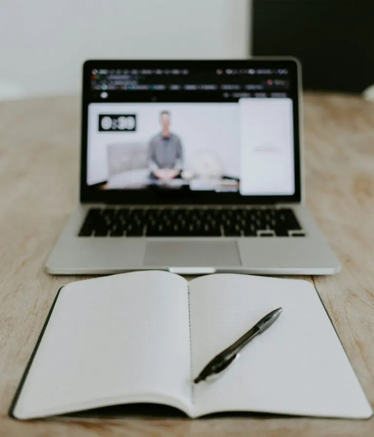 An open notepad and pen is set before a laptop screen with a video running.
