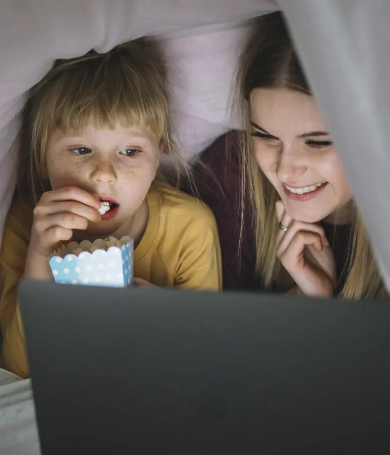 Two sisters, one older and one younger sit under bed sheets watching a movie on a laptop. The younger one eats from a small container of popcorn.