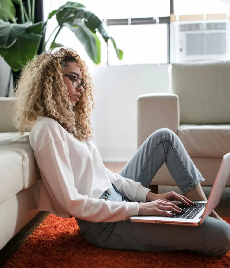 A woman sits on the floor, leaning against a sofa whilst working on their laptop.